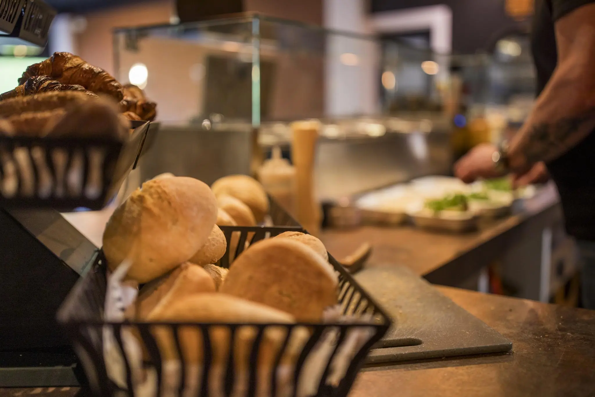 Verse broodjes en croissants in mandjes op een toonbank in lunchroom Markerkant 13 met een medewerker die op de achtergrond voorbereidingen treft.